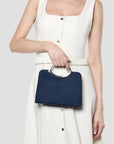 A model wearing a cream-colored sleeveless dress holds a navy blue rectangular handbag with a silver-tone circular handle against a light gray background.