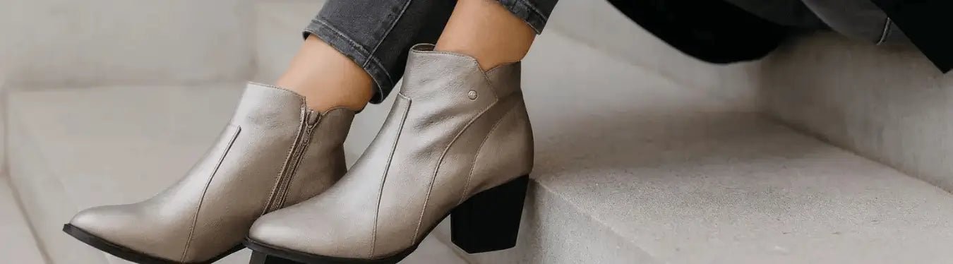 A woman wearing Paradox London metallic pewter wide fit ankle boots sits gracefully on stone steps, showing the boots’ soft sheen and comfortable block heel in a stylish autumn setting.