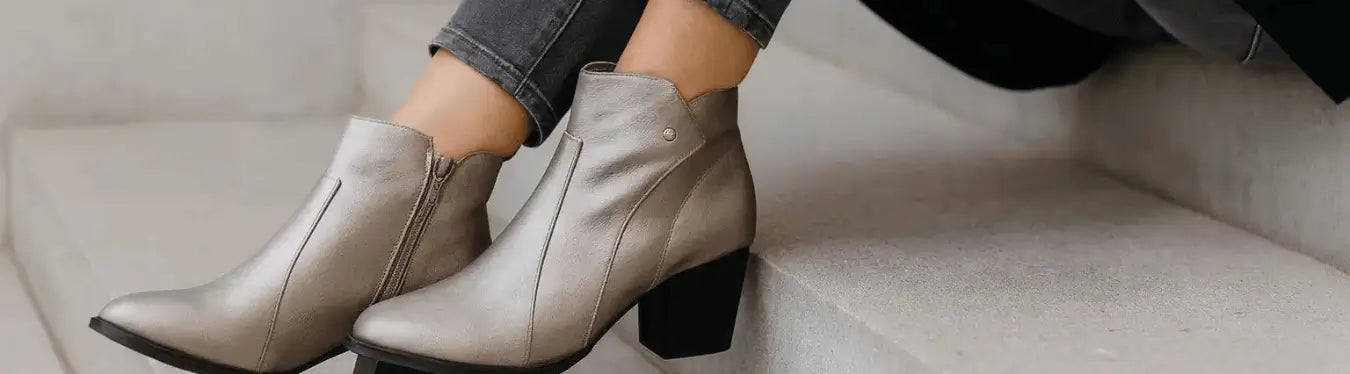 A woman wearing Paradox London metallic pewter wide fit ankle boots sits gracefully on stone steps, showing the boots’ soft sheen and comfortable block heel in a stylish autumn setting.