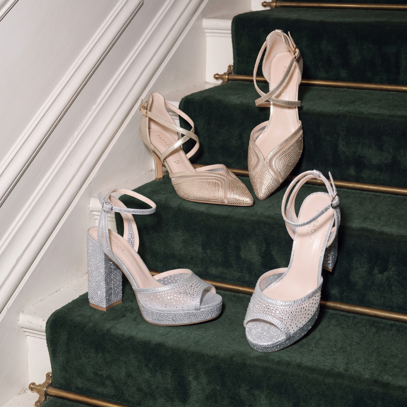 Four women's high-heeled sandals arranged on dark green velvet stairs. Two shoes are metallic champagne gold with pointed toes and thin wraparound straps, and two are silver glitter with open peep-toe platforms and block heels.
