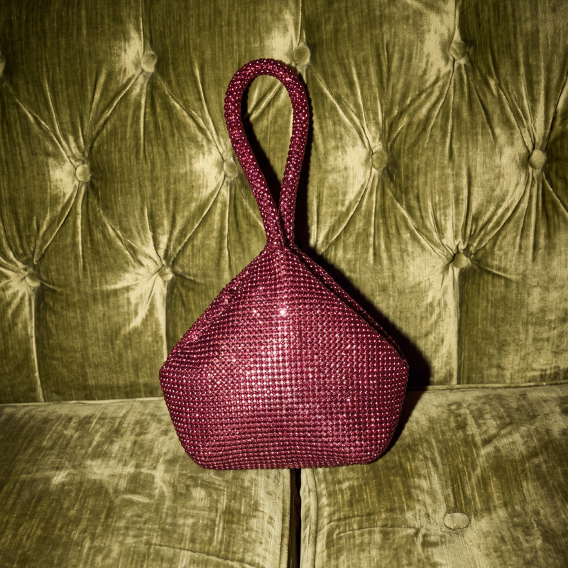 A small structured handbag with a single top handle, covered in sparkling rhinestones in a deep magenta color, displayed on a tufted olive green velvet sofa.
