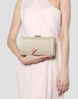 A woman in a pale blush pink draped dress holds a rectangular glitter clutch bag in a metallic champagne gold color with a small gold metal clasp, set against a solid light gray background.