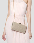 A woman wearing a light blush pink dress carries a small rectangular glitter clutch bag with a gold chain strap against her hip, photographed against a light gray studio background.