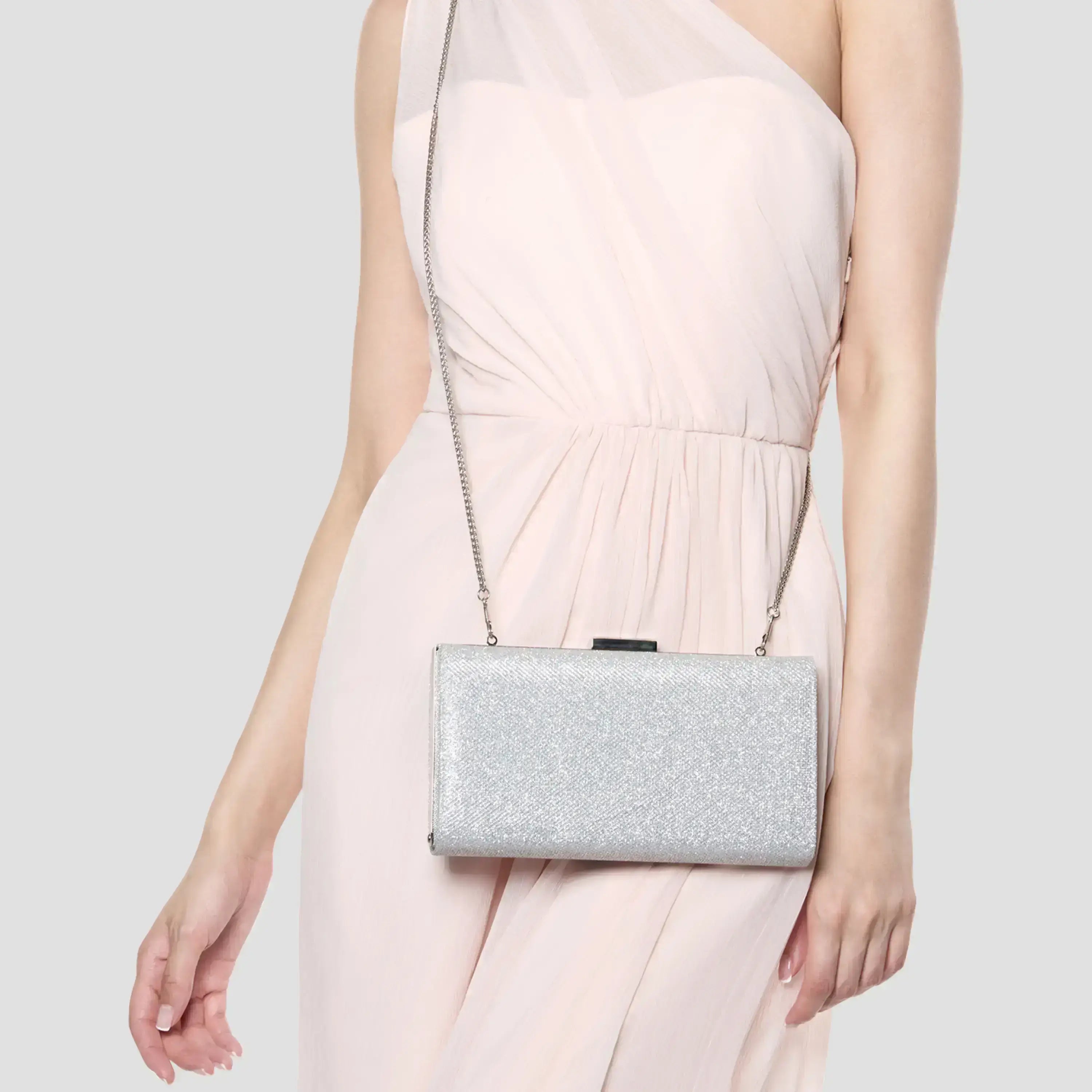 A woman wearing a light pink dress carries a silver glittery rectangular clutch bag with a thin chain strap against a plain light gray background.