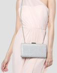A woman wearing a light pink dress carries a silver glittery rectangular clutch bag with a thin chain strap against a plain light gray background.