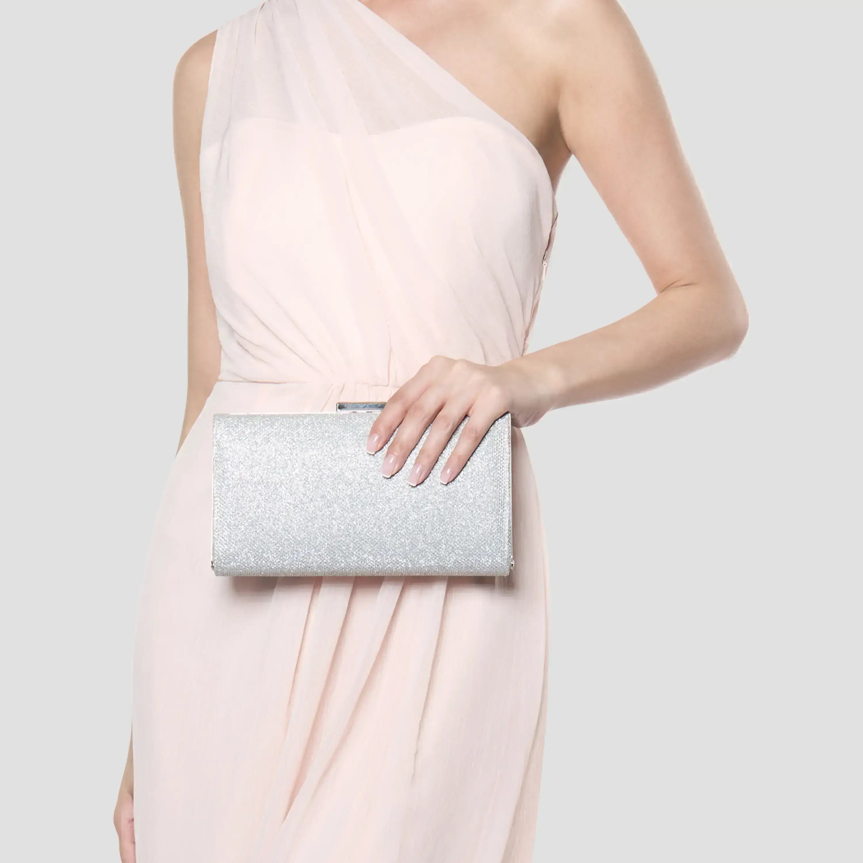 A model wearing a pale blush pink one-shoulder dress holds a silver glitter clutch purse against a light gray studio background.