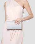 A model wearing a pale blush pink one-shoulder dress holds a silver glitter clutch purse against a light gray studio background.