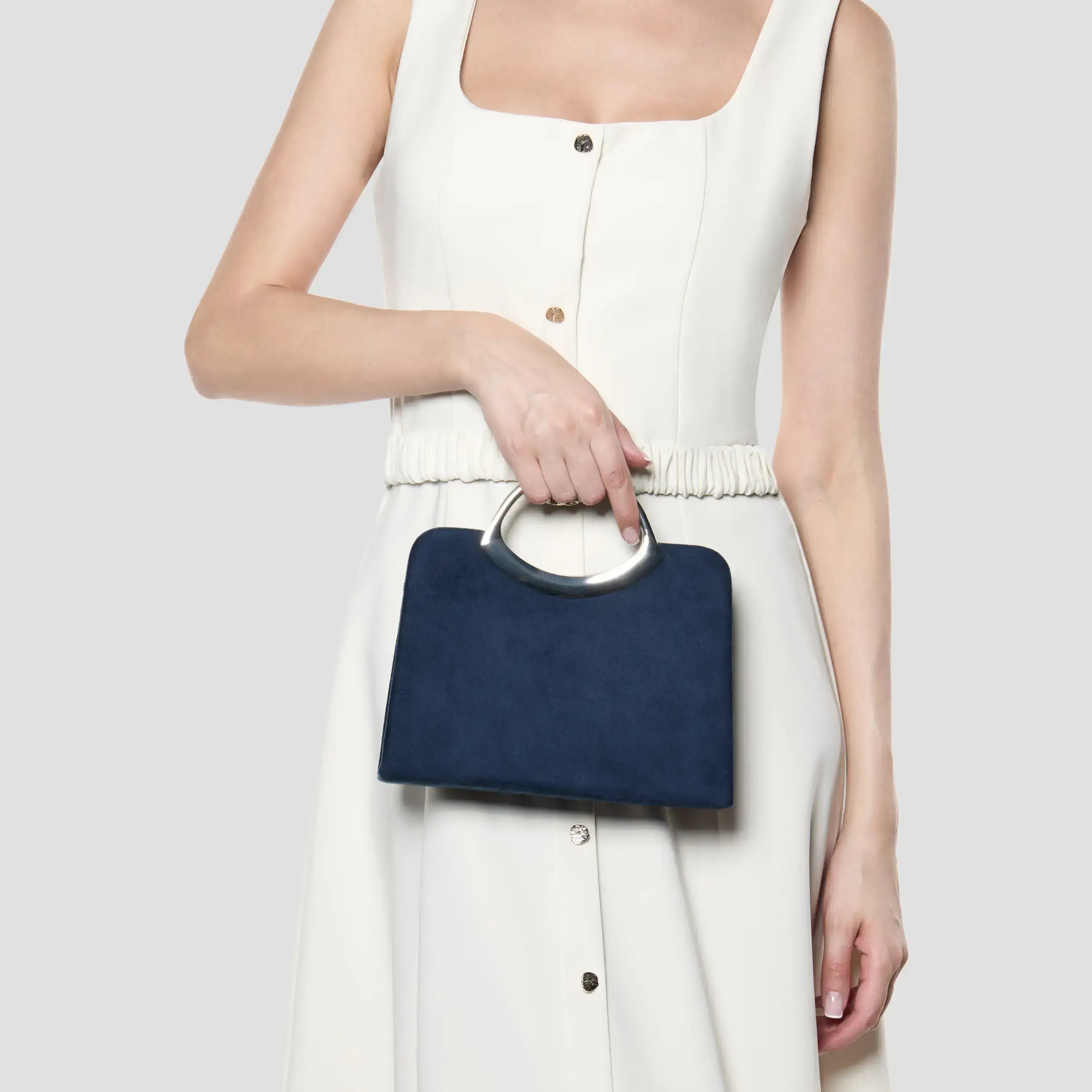 A model wearing a cream-colored sleeveless dress holds a navy blue rectangular handbag with a silver-tone circular handle against a light gray background.