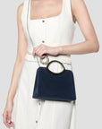 A navy blue velvet handbag with a circular black handle and silver chain strap, held by a model wearing a cream-colored sleeveless dress against a light gray background.