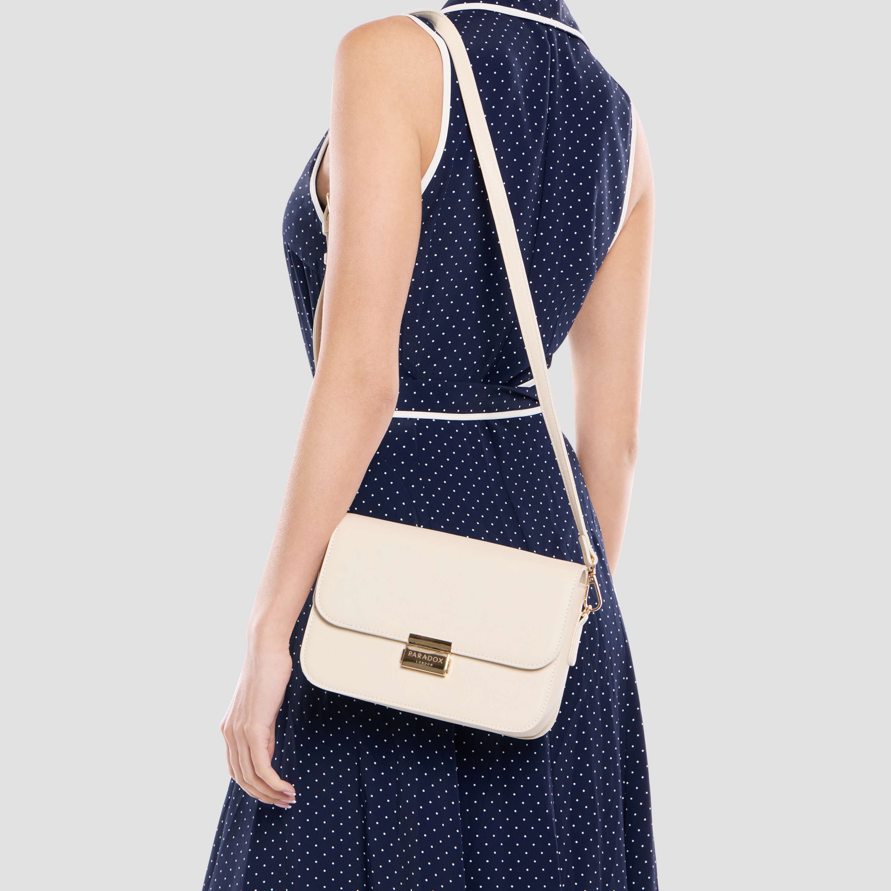 A woman wearing a navy blue and white polka dot dress is shown from the back, carrying a cream-colored crossbody bag with a gold-tone clasp against a light gray background.