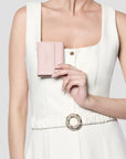 A model in a white sleeveless dress holds a small blush pink leather clutch purse with gold hardware against her chest.