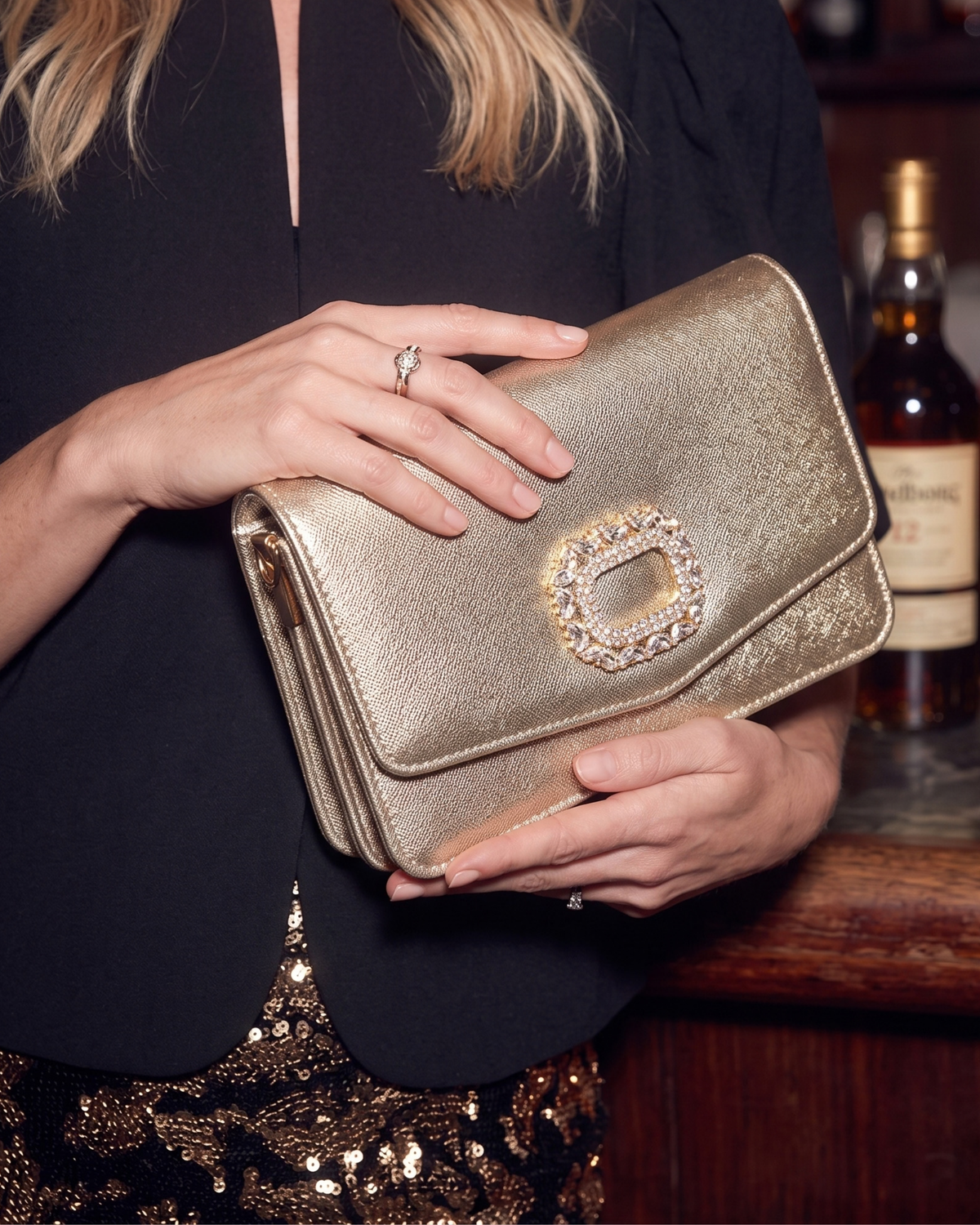A woman in a black blazer and sequined skirt holds a metallic gold envelope-style clutch bag with a jeweled square clasp. A bar setting with a bottle is visible in the background.
